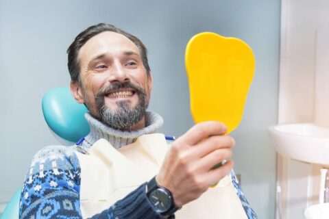 Mann hält einen Spiegel in der Hand und begutachtet seine Zähne